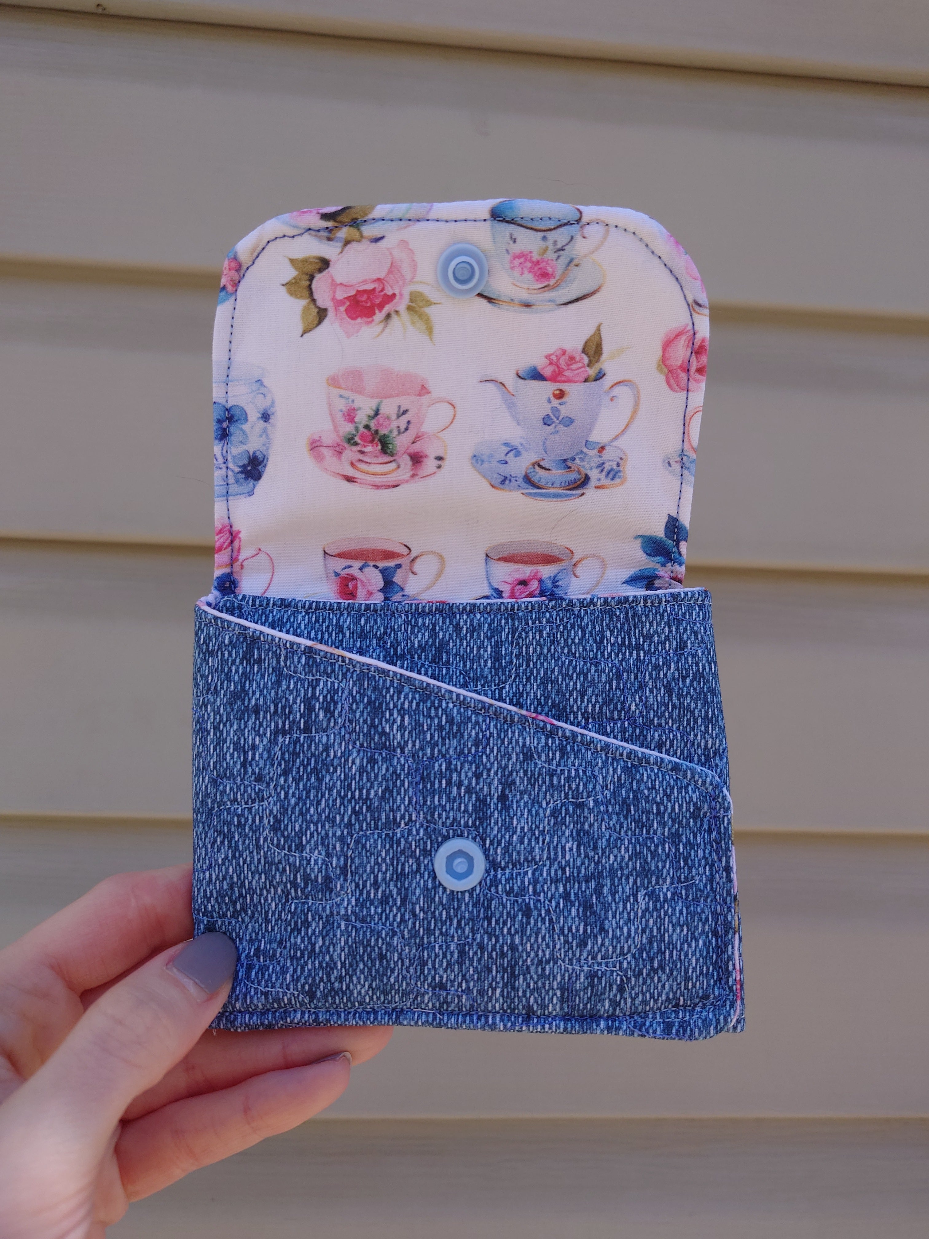 A handmade fabric wallet with a floral design featuring teapots, predominantly in blue and white colors, held in a person's hand.