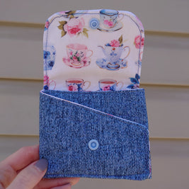 A handmade fabric wallet with a floral design featuring teapots, predominantly in blue and white colors, held in a person's hand.