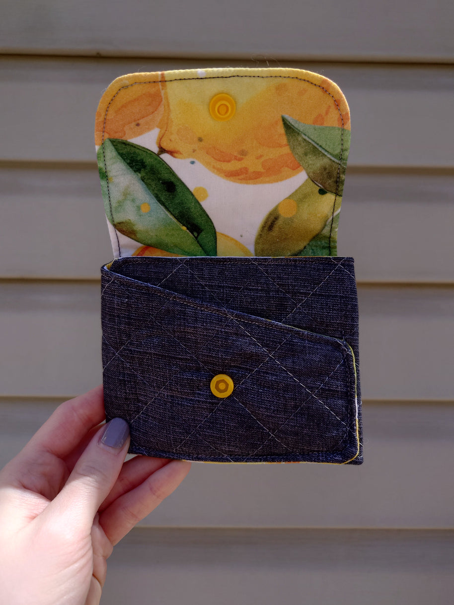 A handmade fabric wallet with a lemon print on the top compartment and a solid blue color on the bottom. It features a fold-over design with a visible yellow snap button.