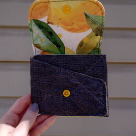 A handmade fabric wallet with a lemon print on the top compartment and a solid blue color on the bottom. It features a fold-over design with a visible yellow snap button.