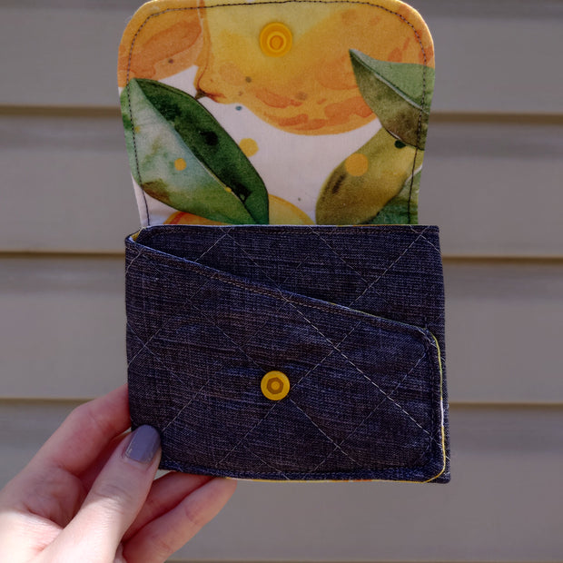 A handmade fabric wallet with a lemon print on the top compartment and a solid blue color on the bottom. It features a fold-over design with a visible yellow snap button.